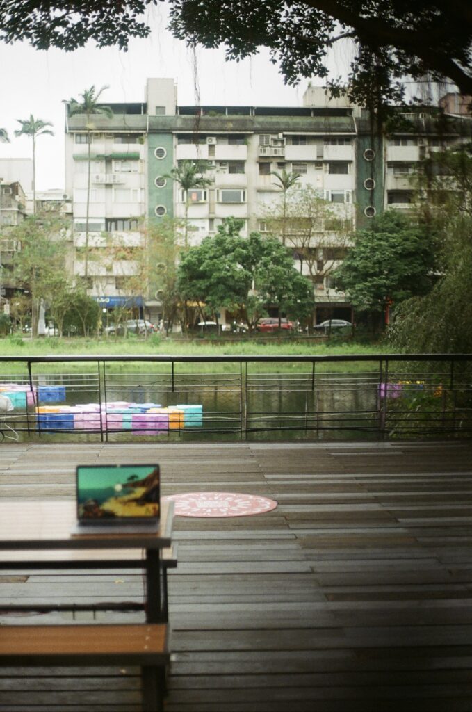 Image is a scanned film picture showing a colorful scene at Songshan Cultural and Creative Park: a bench where I sat to work, the pond, colorful art installation in the water, and green trees.